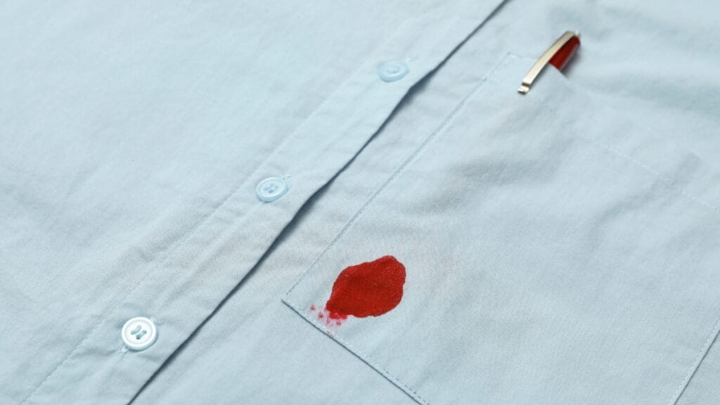 White fabric shirt laid flat with a visible red blood-like stain near the center, next to a loose thread and surrounded by visible white stitching buttons and seams, “how to get blood out of clothes”.