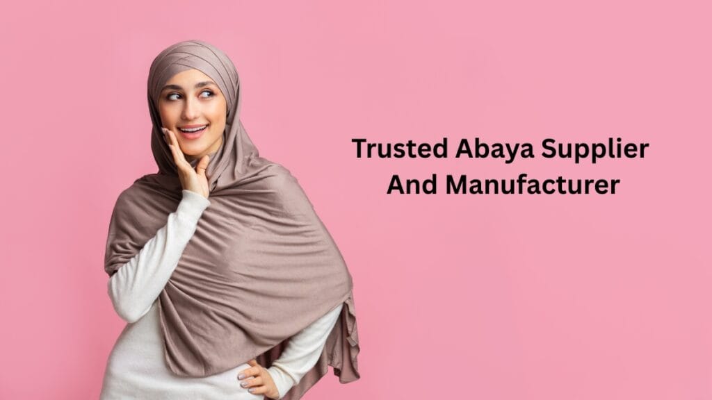 A smiling woman wearing a modest abaya and a taupe hijab stands against a pink background, looking to the side with her hand near her face. Text on the image reads: “Trusted Abaya Supplier And Manufacturer.”