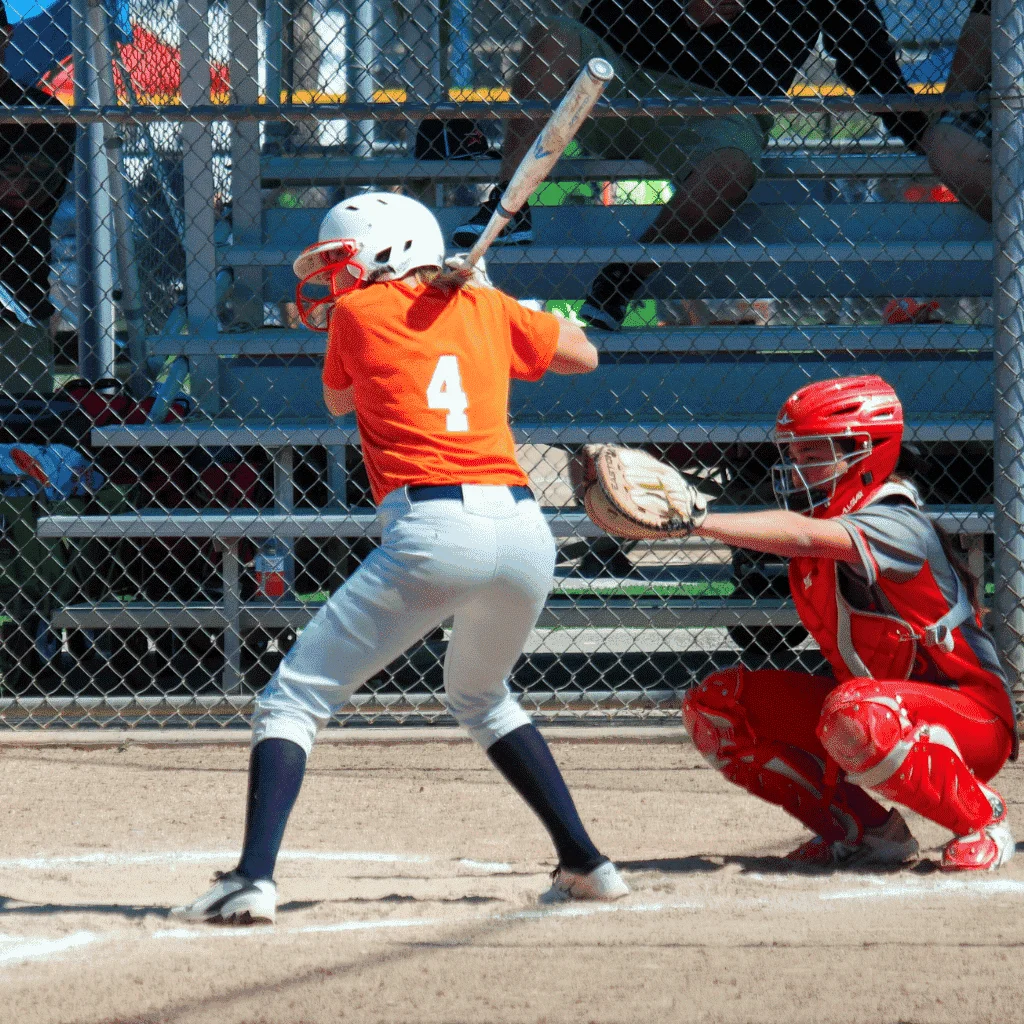 Exciting softball Uniforms by Fashion Soul International, featuring a batter in an orange jersey swinging at a pitch, with a catcher in red gear behind home plate.