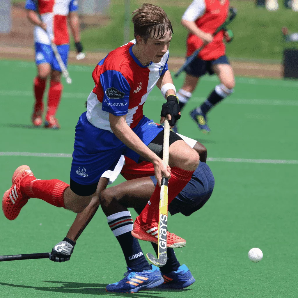 Field hockey player in red and blue uniform controlling the ball with a Grays stick, with another player in the background.