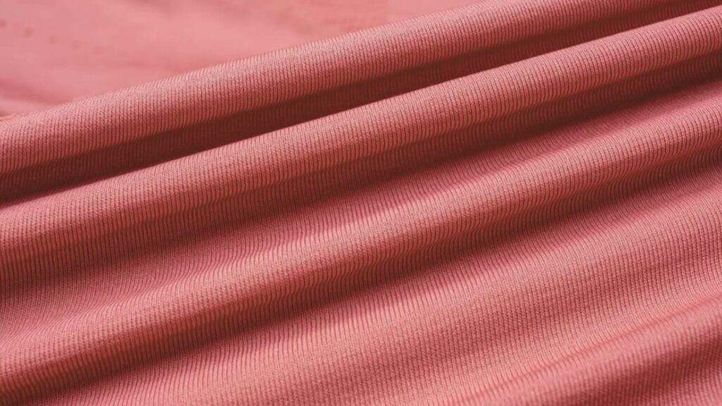  A close-up view of coral-colored ribbed fabric with soft folds and wrinkles, showing the knit texture and gentle undulations in the material.
