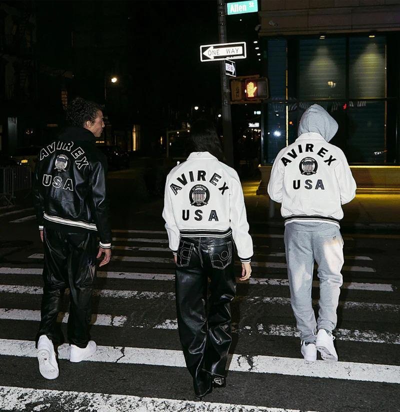"Three stylish individuals wearing AVIREX USA jackets walking at night in an urban setting. The left person is dressed in a black leather jacket and pants, the center person in a white bomber jacket with black pants, and the right person in a gray hoodie and sweatpants. They are crossing a street with streetlights and a one-way sign visible."