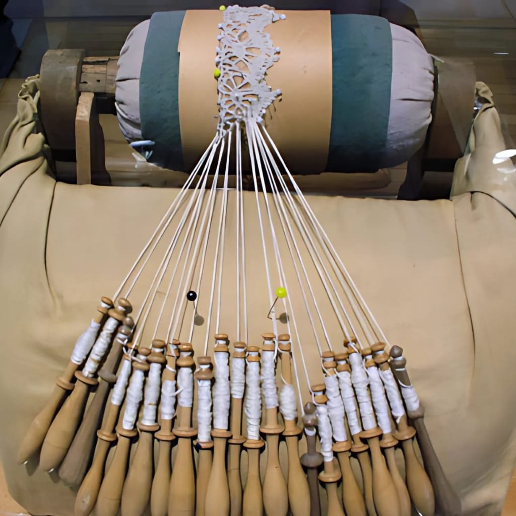 A collection of wooden badminton rackets with white grip tape, laid out on a beige cloth surface. The rackets are arranged in a semi-circle, with some pins sticking through the grips. In the background, there is a wooden table with a decorative white lace doily on a rolled-up cushion.