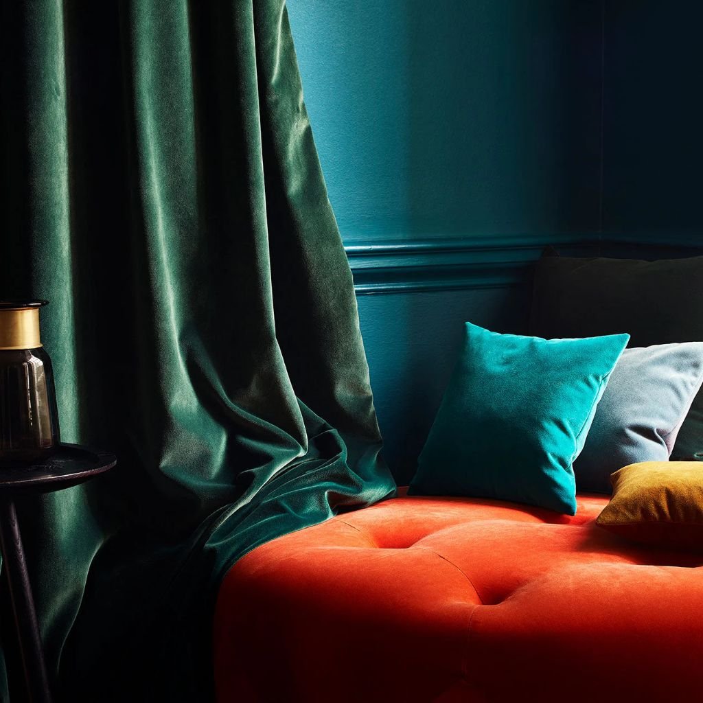 A cozy interior corner featuring a deep orange tufted cushion or sofa, with dark teal curtains draped to the side, set against a blue-green wall. There are two pillows—one teal and one yellow—resting on the cushion, and a small black side table with a dark glass jar or lantern on it.