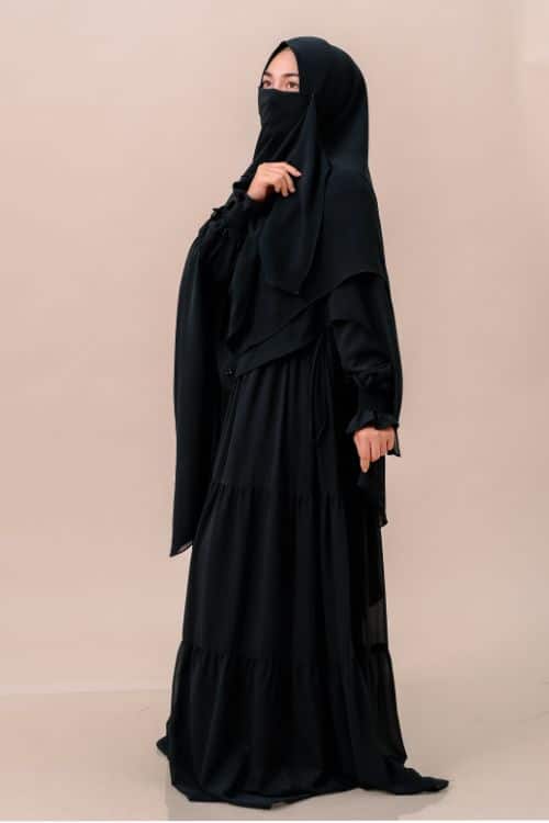 A woman wearing a black hijab and a tiered black dress poses elegantly against a neutral background, exemplifying modest fashion.