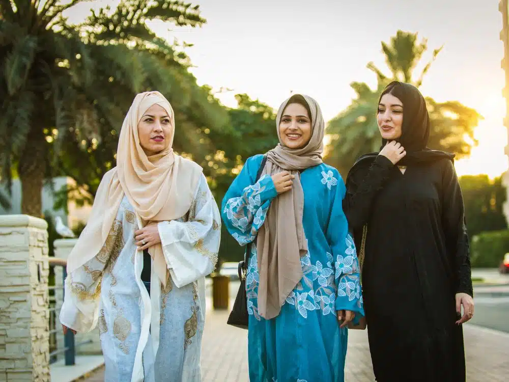 Three women in traditional outfits walking together outdoors during sunset, surrounded by palm trees.
