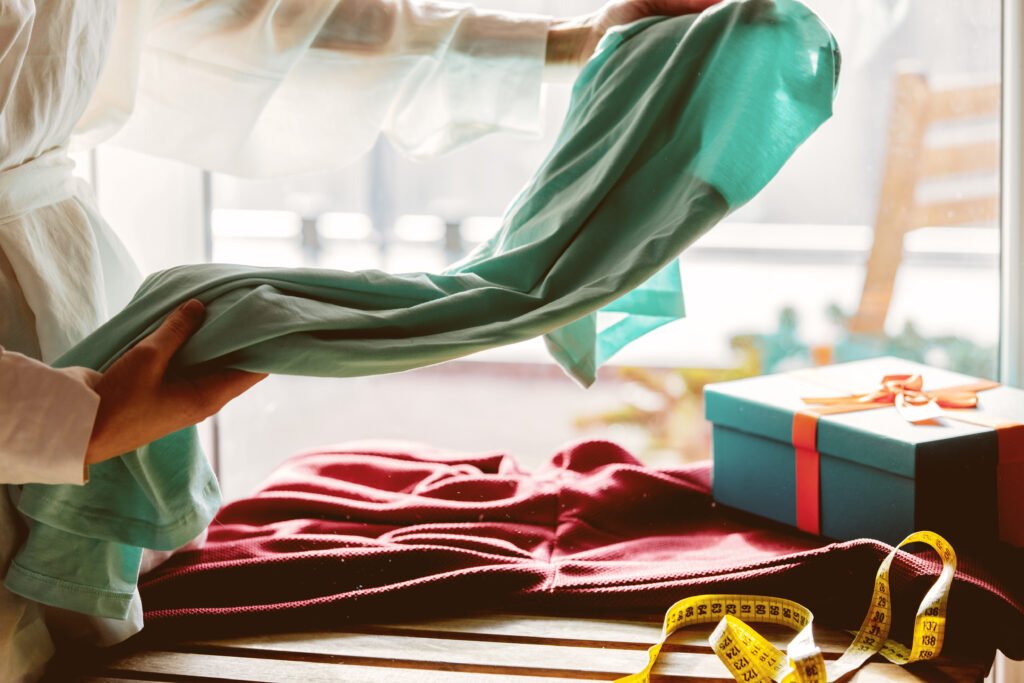 A person is holding a piece of turquoise fabric above a red garment on a wooden table. In the background, there is a wrapped gift box with a ribbon and a measuring tape.