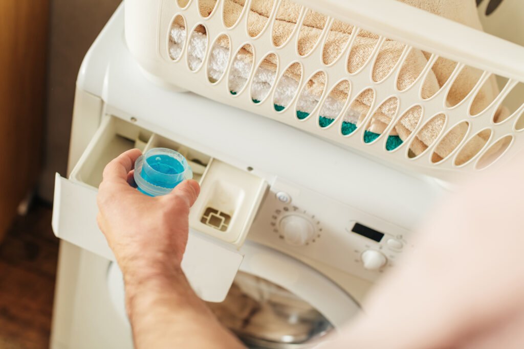 "A person holding a blue laundry detergent container near the detergent drawer of a washing machine, with a laundry basket of towels in the background." Smart & safely