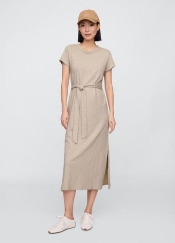 A woman wearing a belted tee dress in a light beige color, paired with a tan cap and white sneakers. The dress features short sleeves, a round neckline, and a tied belt at the waist.