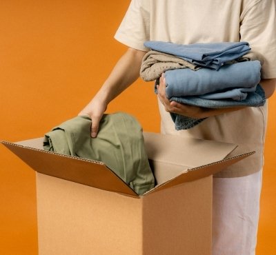 A person is placing folded clothes into a cardboard box against an orange background.