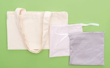 A collection of cotton bags, including a large beige tote bag and two smaller pouches in light pink and gray, situated against a green background.