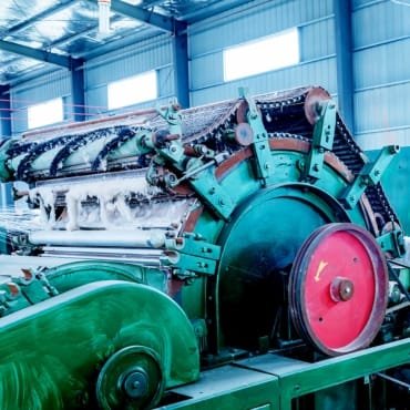 A large, industrial cotton gin machine inside a well-lit facility, featuring a complex array of gears, belts, and cotton fibers circulating through the mechanism.