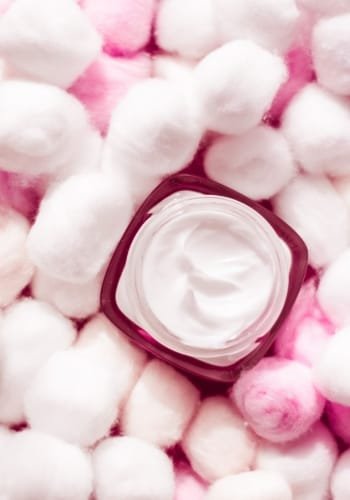 A close-up of a jar of cream surrounded by fluffy cotton balls in pink and white.