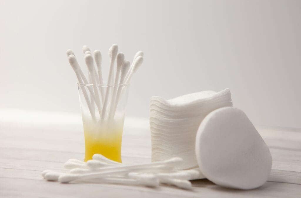 A glass filled with cotton swabs next to a stack of white cotton pads on a light-colored surface.
