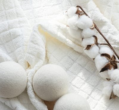 Three white wool dryer balls are placed on a soft, textured white towel, alongside a sprig of cotton plants with fluffy white bolls. 
