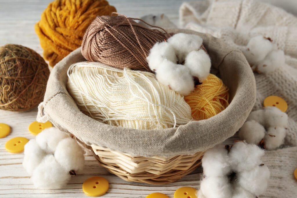 A cozy basket filled with various skeins of yarn in yellow, brown, and white, surrounded by cotton bolls and yellow buttons on a wooden surface.