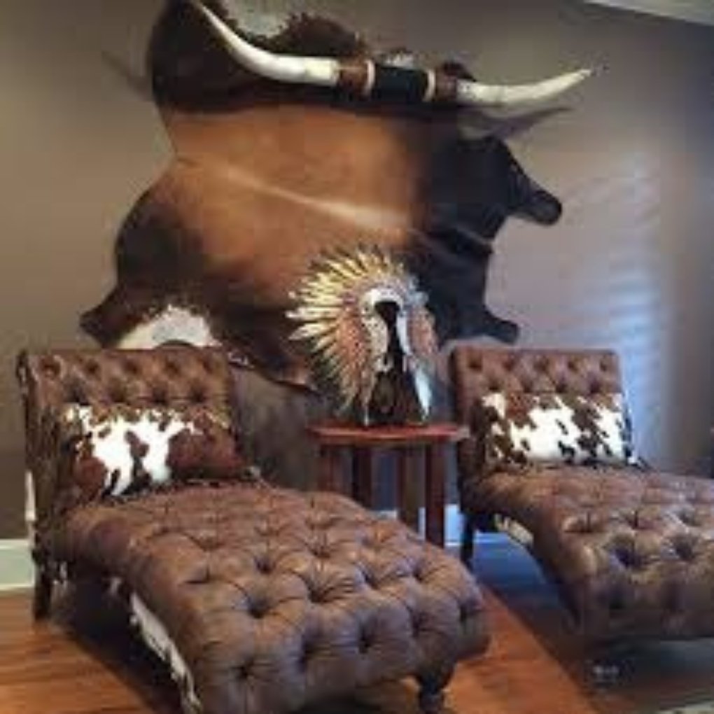 A rustic sitting room featuring two brown tufted chaise lounges, a wooden side table, and a decorative Native American headdress mounted on the wall, alongside a large bullhide rug.