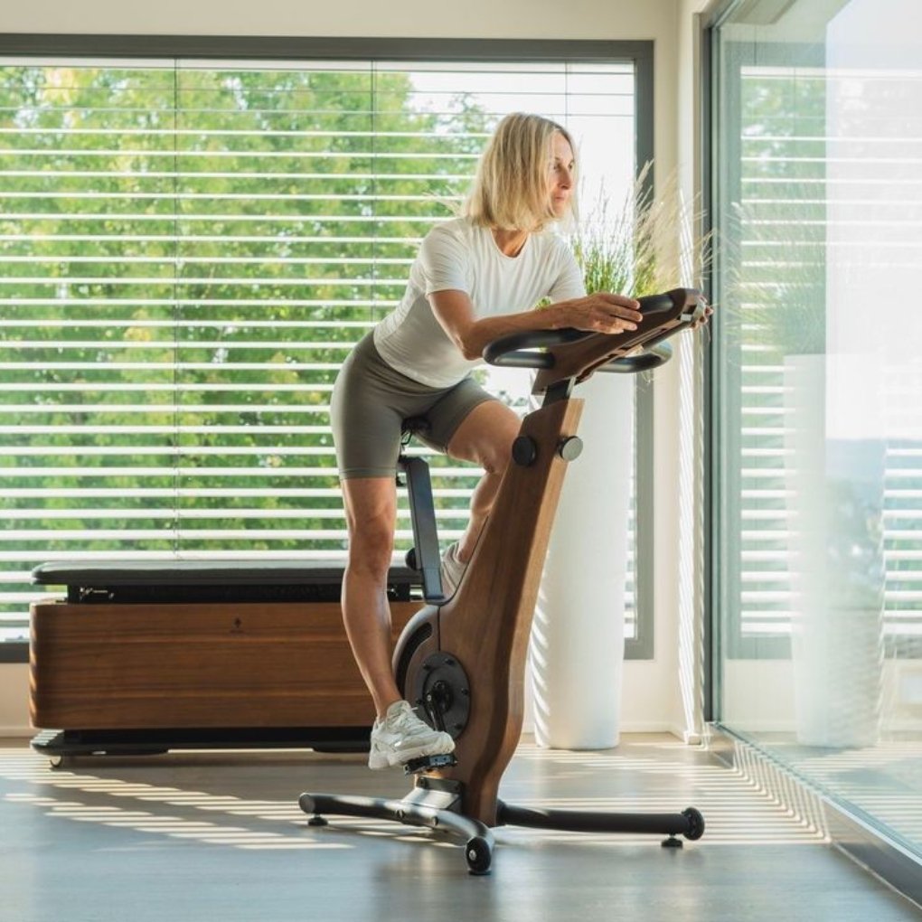 A woman with blonde hair is exercising on a modern wooden stationary bike in a bright room with large windows and blinds. She is dressed in a white shirt and gray shorts, focusing intently on her workout.
Gym wear guid