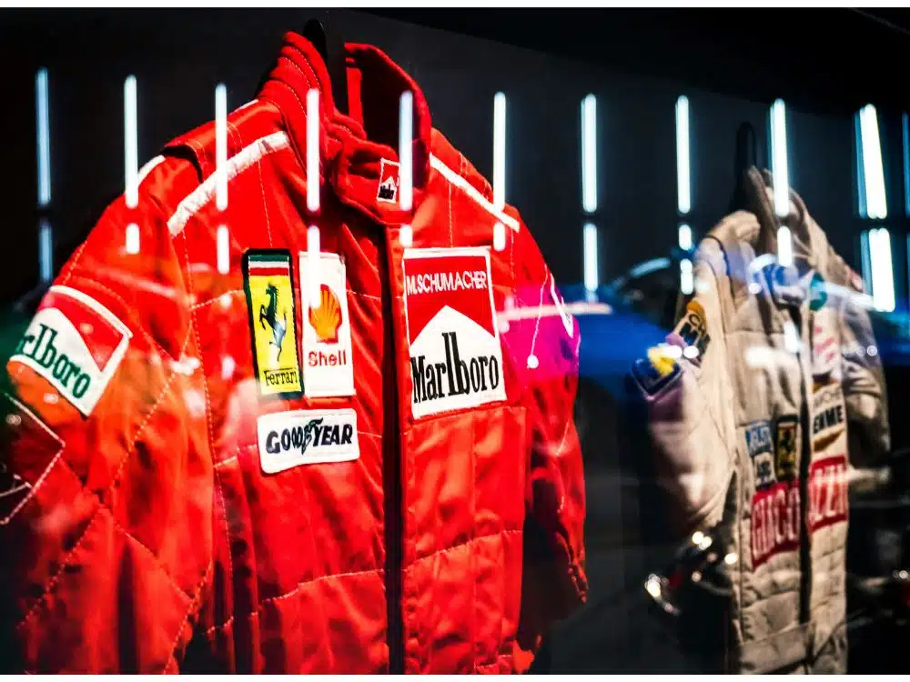 Display of iconic racing suits, including a red Ferrari suit and a white suit, in a showcase.