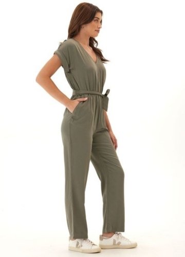 A woman stands confidently in a relaxed olive green jumpsuit with short sleeves, a V-neckline, and a cinched waist. She wears white sneakers and poses with her hands in the jumpsuit's pockets.