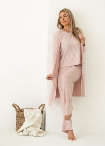 A woman in a pink knitted lounge outfit, styled with a long cardigan, poses casually. There’s a woven basket beside her.