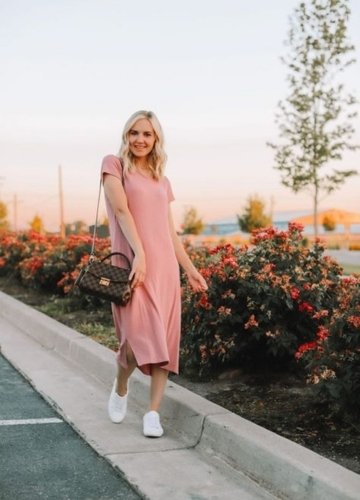 A woman wearing a pink tee dress with white sneakers, walking along a paved path lined with blooming flowers in a serene outdoor setting during sunset.