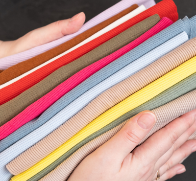 A person holding a stack of colorful ribbed fabric materials, including pink, blue, yellow, brown, and green.