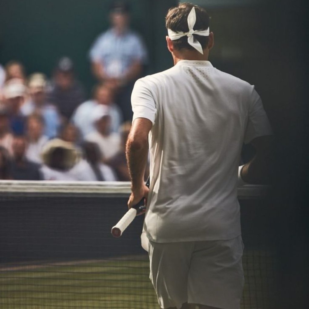 A tennis player in a white outfit and headband stands with a racket in hand, facing a crowd during a match, with a focus on his back.