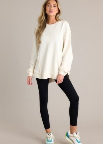 A model wearing an oversized cream sweatshirt, black leggings, and colorful sneakers, posing against a neutral background.