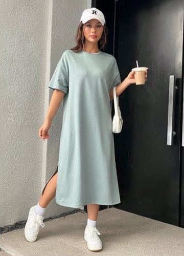 A woman wearing an oversized mint green tee dress with side slits, holding a cup of coffee, and wearing a white cap and sneakers. 