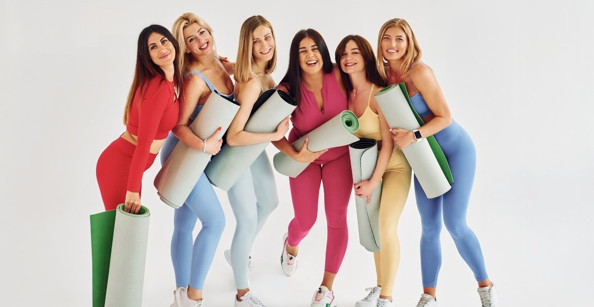 A group of six women in colorful athletic wear, each holding yoga mats, posing together and smiling joyfully.