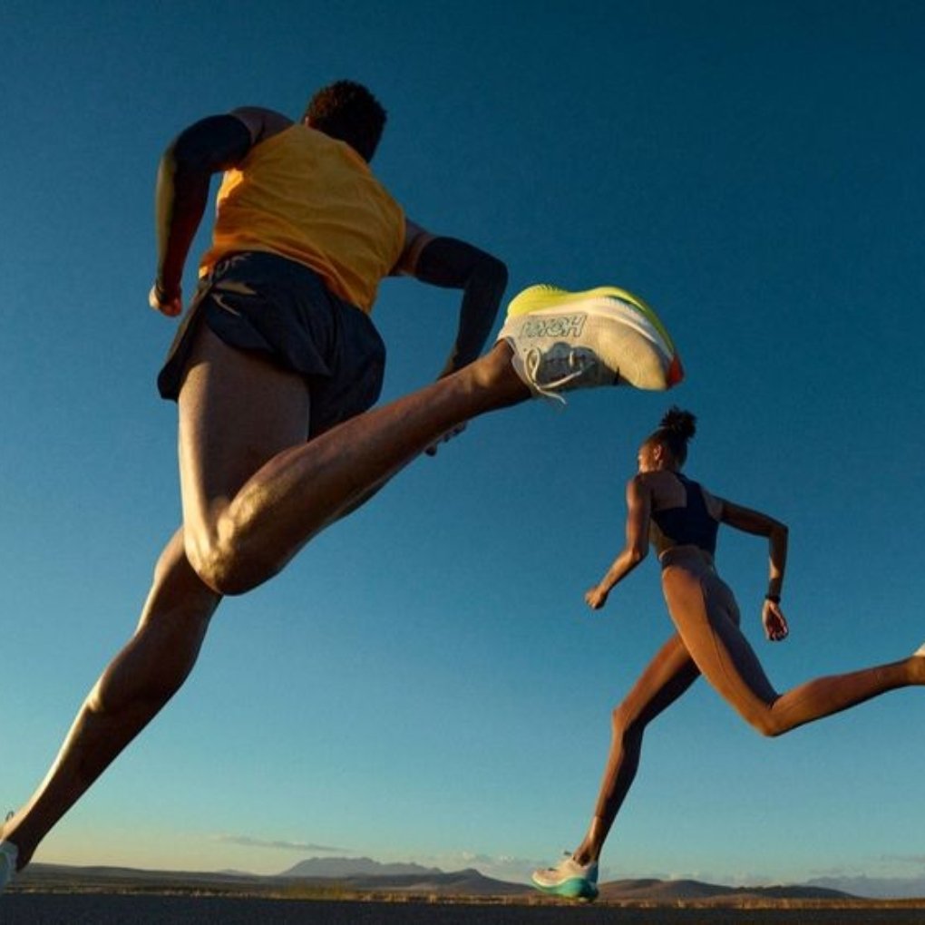 Two runners in athletic gear sprinting on a road against a clear blue sky during sunset, highlighting their movement and powerful strides.