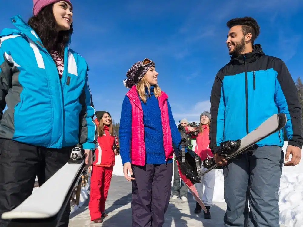 Group of young people in winter clothing smiling and walking with skis in a snowy outdoor setting.