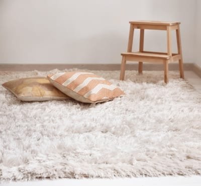 A cozy room featuring a plush white rug with two decorative pillows—one with a striped pattern and the other with a geometric design. A small wooden step stool is in the background.