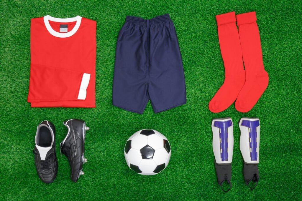 A flat lay image of soccer gear on green grass. Items include a red jersey, navy shorts, red socks, black soccer cleats, a black and white soccer ball, and white shin guards with purple accents. products from the cotton