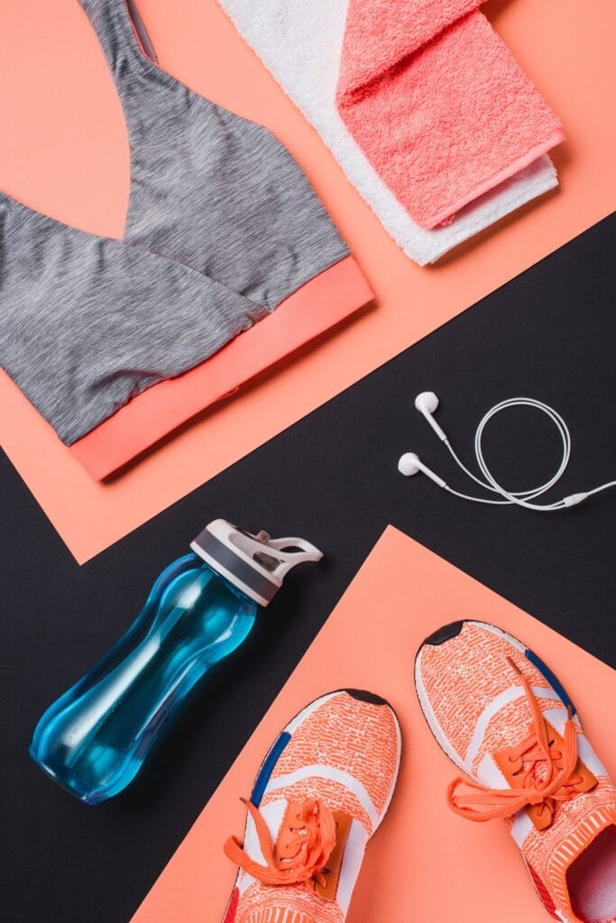 A flat lay arrangement of sports and fitness items, including a gray sports bra, two towels (one white, one coral), a blue water bottle, orange athletic shoes, and white earphones, all on a colored background.