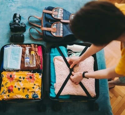 Person packing a suitcase with clothing and travel essentials such as a camera, toiletries, and face masks.