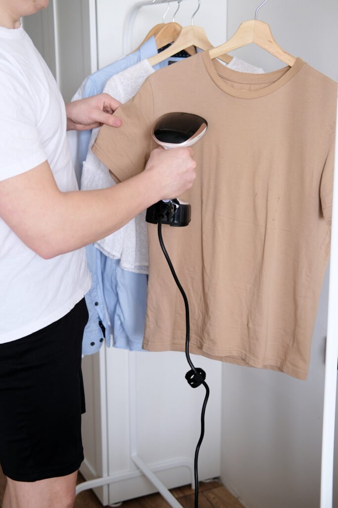 A person using a clothes steamer on a brown t-shirt hanging on a rack, with other shirts in the background.