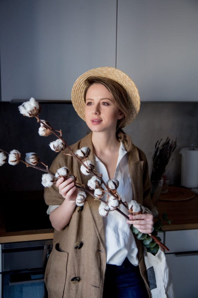 A woman holding a branch of cotton while wearing a straw hat and a loose, light-colored outfit, standing in a modern kitchen.
