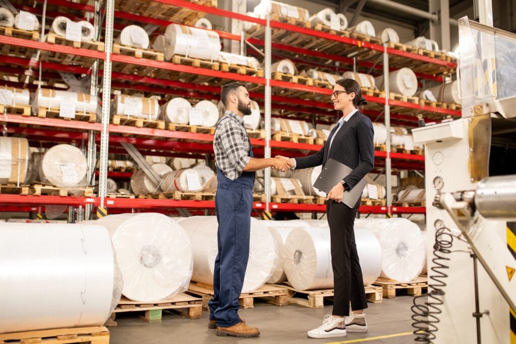 A man and a woman shake hands in a warehouse filled with rolls of paper on pallets. The man is wearing a checkered shirt and overalls, while the woman is dressed in formal business attire.