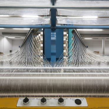 Close-up of a cotton weaving machine in a textile factory, showing multiple spools of yarn aligned horizontally and threads being woven into fabric, with spindles and guiding mechanisms visible.
