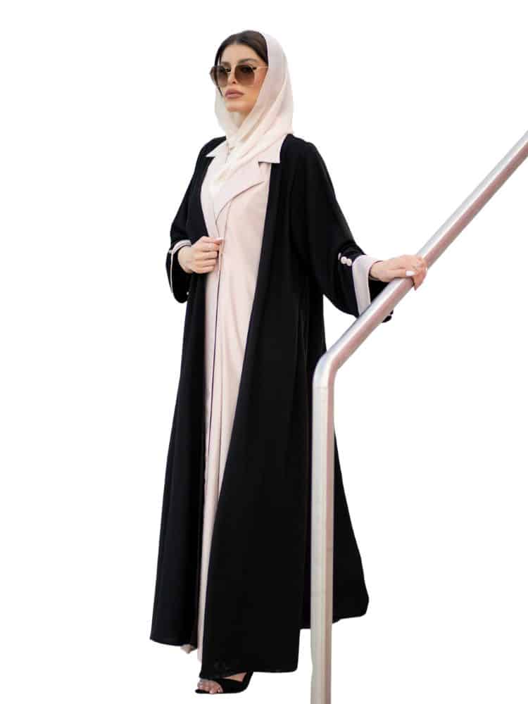 A woman in a stylish black abaya and a light-colored hijab stands on a staircase, wearing sunglasses and holding a banister.