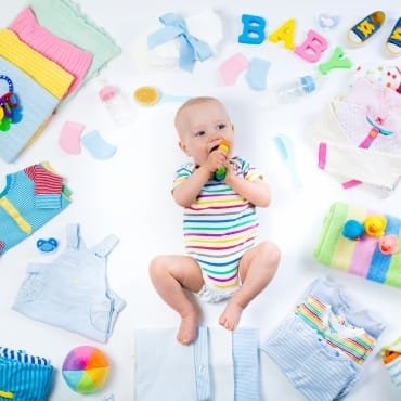 A smiling baby wearing a striped onesie, surrounded by colorful cotton baby clothes, toys, and accessories.
