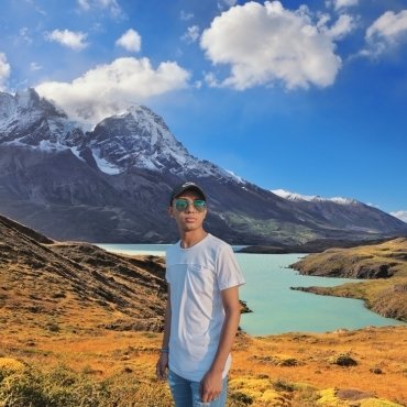 A young man standing in front of a scenic mountain landscape with a lake, wearing a white summer t-shirt and sunglasses.