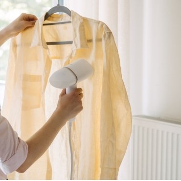 A person using a hairdryer to remove wrinkles from a hanging light-colored rayon shirt.
