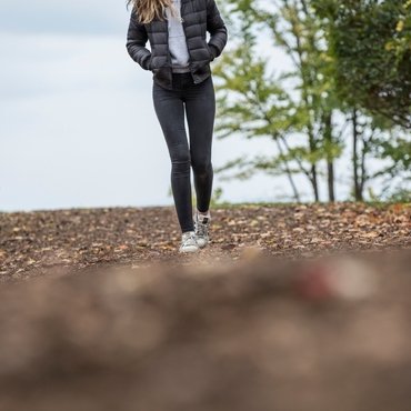 A woman walking along a trail wearing a puffer jacket, a gray hoodie, and black leggings with sneakers.