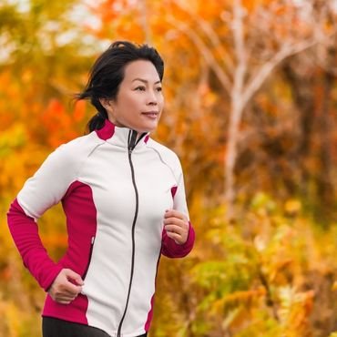 A woman jogging outdoors in a white and pink zip-up jacket amid colorful autumn foliage.