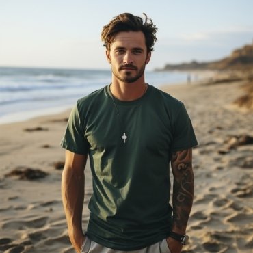 A young man with tousled hair wearing a green t-shirt and a necklace stands confidently on a sandy beach with waves in the background at sunset.