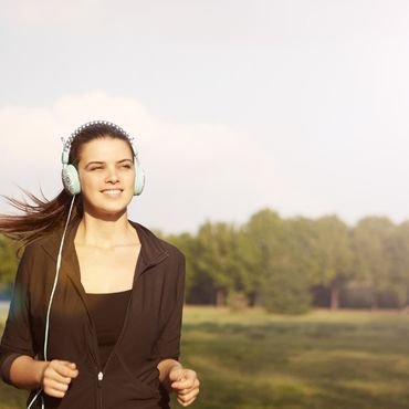 A woman running outdoors while wearing headphones and a black zip-up jacket, smiling as she enjoys her trendy workout outfits.