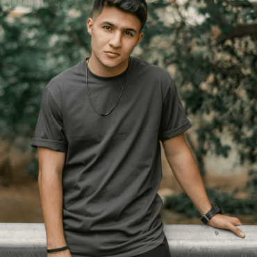 A young man wearing a dark gray summer t-shirt and a necklace poses casually against a blurred outdoor background.